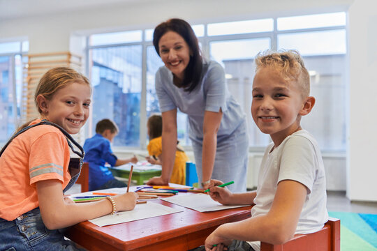 Creative kids during an art class in a daycare center or elementary school classroom drawing with female teacher.