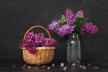 Wicker basket and glass vase of beautiful lilac flowers on table near grunge black wall