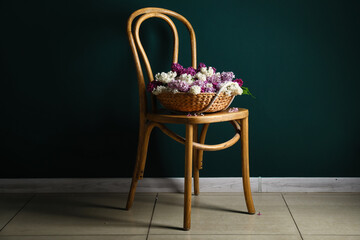 Fototapeta na wymiar Chair with wicker basket of beautiful fragrant lilac flowers near dark green wall
