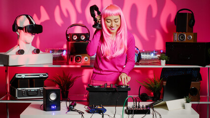 Happy artist mixing electronic remix at professional mixer console, listening music into headphones during party. Asian dj performer with pink hair dancing and having fun in club at night.