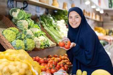 Delighted young Muslim woman purchaser choosing red tomatos at the counter in grocery store