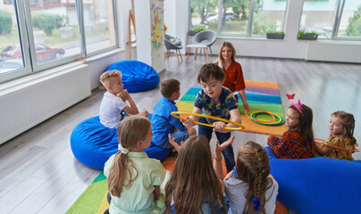 A happy female teacher sitting and playing hand games with a group of little schoolchildren - obrazy, fototapety, plakaty