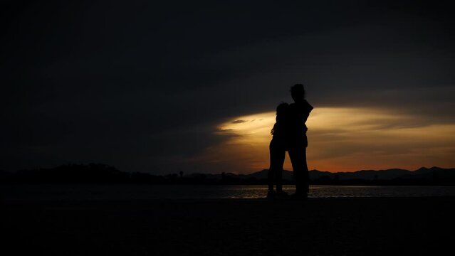 Mum and child silhouette on tropical sunset. Woman with her child silhouette stay on the shore and admire the colorful dark sky in summer. A concept of family vacation and free time.