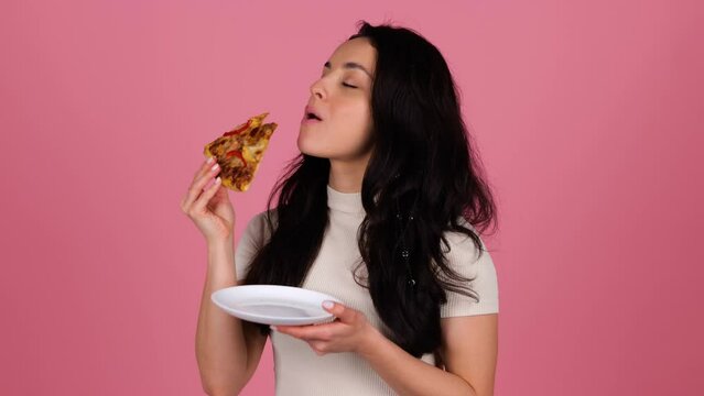 Smiling beautiful female biting pizza while standing and posing in studio over pink background. People, lifestyle, junk food concept. Real time