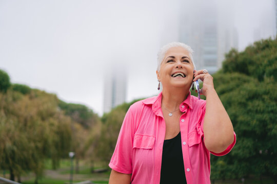 Older Gray Haired Latin Woman Talks On Cell Phone While Laughing Outside, Copy Space