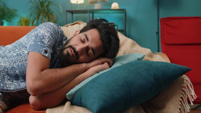 Tired indian man lying down in bed taking a rest. Carefree young hindu guy napping, falling asleep on comfortable sofa with pillows at home apartment room. Closed his eyes enjoy daytime nap alone