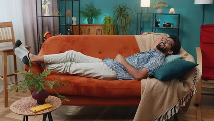 Tired indian man lying down on comfortable couch, taking a rest, daytime nap. Portrait of carefree hindu guy holding hands behind head relaxing, falling asleep at home apartment living room on sofa