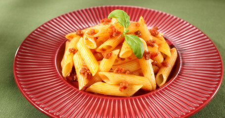 pasta bolognese. penne rigato with ragout sauce. Italian cuisine dish with tomato meat sauce and basil leaf. red plate with food on a green background. angle view. close-up.