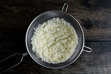 Thawed Frozen Cauliflower Rice in a Mesh Strainer: Rice cauliflower in a colander placed over a mixing bowl
