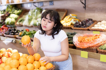 Obraz premium Pensive young asian girl selecting ripe oranges while shopping in fruits and vegetables section of supermarket..