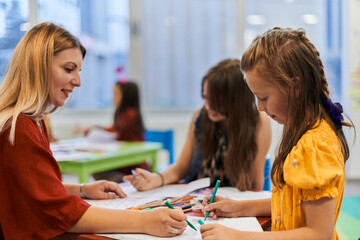 Creative kids during an art class in a daycare center or elementary school classroom drawing with female teacher.