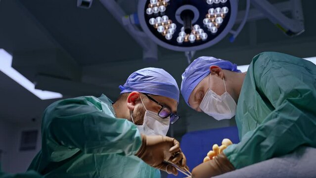 Bearded surgeon uses forceps carefully performing operation on foot. Assisting doctor supports the operated limb. Low angle view.