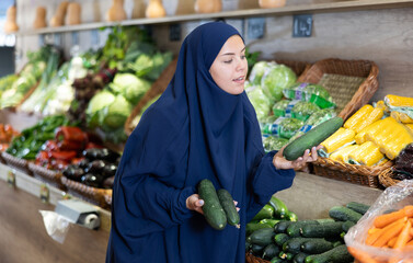 Delighted young Muslim woman purchaser choosing fresh cucumbers at the counter in grocery store