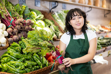 Smiling young saleswoman holding gladly beetroots at the counter in big grocery store