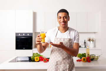 young african american man in apron holds glass with fresh orange juice and smiles, the guy advertises healthy food