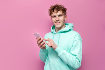 young curly guy in mint hoodie uses smartphone on pink isolated background, man in blue sweatshirt...