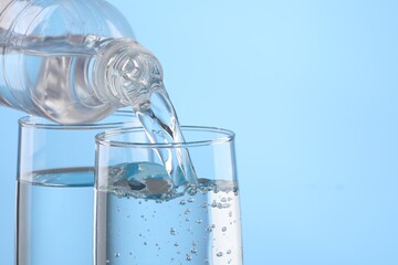 Pouring water from bottle into glass on light blue background, closeup. Space for text