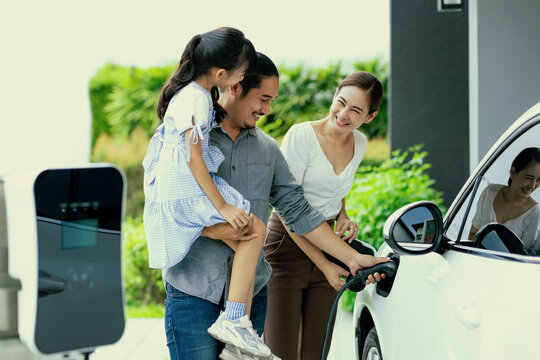 Progressive young parent teach daughter how to recharge or refuel EV car at home charging station. Green and clean energy from electric vehicle for healthy environment. Eco power from renewable source
