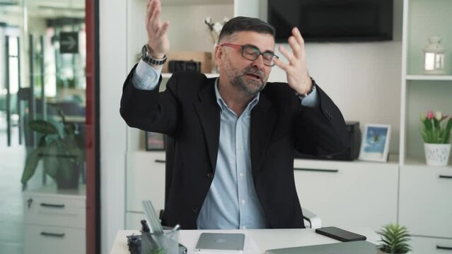 A man in a business suit is sitting at a desk in an office and arguing with someone off-camera, showing confusion and dissatisfaction.
