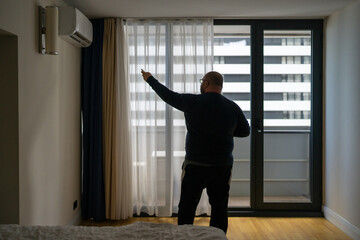 Overweight man switching on air conditioner at home in summer using remote control. Silhouette male suffers from heat stuffiness in hotel room. Overheated man feels abnormally high air temperature.