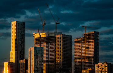 High-rise residential buildings under construction along with construction cranes illuminated by the setting sun against a dark blue sky