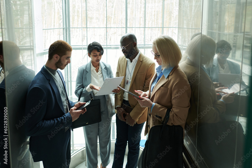 Leinwandbilder group of intercultural employees looking through points of their reports while moving in elevator an