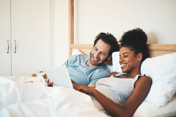 Happy couple, tablet and relax on bed for morning entertainment or online streaming together at home. Interracial man and woman person relaxing in bedroom on technology for social media or browsing