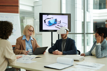 Young man with vr headset sitting by table among colleagues during presentation at meeting while mature woman using laptop