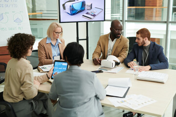 Several intercultural coworkers in formalwear discussing working points and networking while sitting by table in boardroom or office