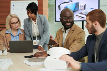 Happy young African American businessman talking to colleague with vr headset against two women discussing working points at meeting