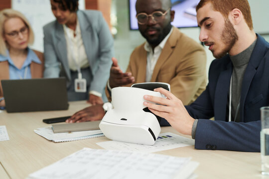 Focus on young businessman making presentation of vr headset to African American male colleague while sitting against two businesswomen - Powered by Adobe