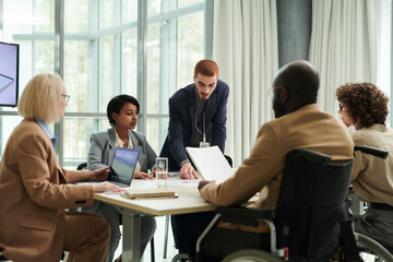 Young confident chief executive officer pointing at document on desk while explaining financial or graphic data to group of economists