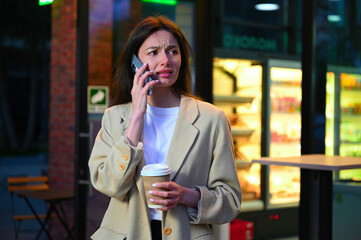 A beautiful woman dressed in casual attire is enjoying a cup of coffee in an urban setting. She appears to be deep in conversation as she speaks on her phone, with an expression