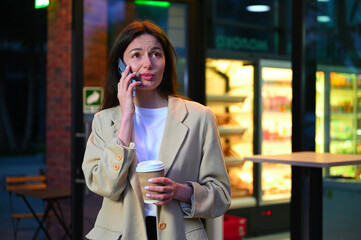 A beautiful woman dressed in casual attire is enjoying a cup of coffee in an urban setting. She appears to be deep in conversation as she speaks on her phone, with an expression