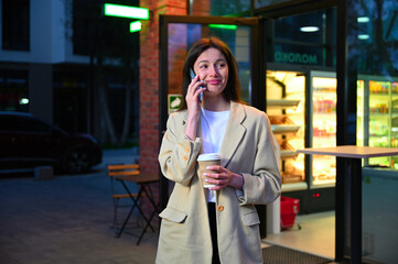 Fototapeta premium A beautiful woman dressed in casual attire is enjoying a cup of coffee in an urban setting. She appears to be deep in conversation as she speaks on her phone, with an expression