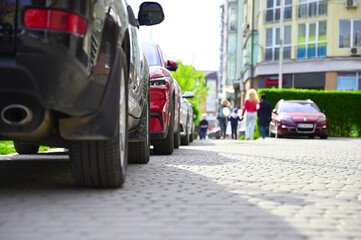 A close up shot of the wheels of a car in urban parking lot, with people and other vehicles visible in the background. The focus is on the textureand detail of the tires.