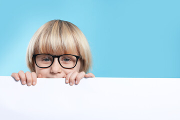 Cute little boy in glasses with blank board on light blue background, space for text