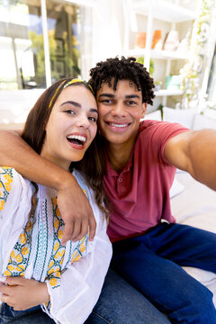 Portrait of happy diverse couple smiling for selfie and embracing, sitting in living room at home