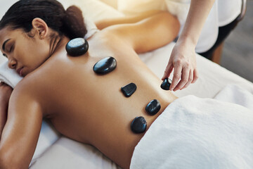 Woman, hands and stone massage at spa for skincare, beauty or body treatment on bed at resort. Calm female lying in relaxation with masseuse, hot rocks and back for therapy, zen or wellness at salon