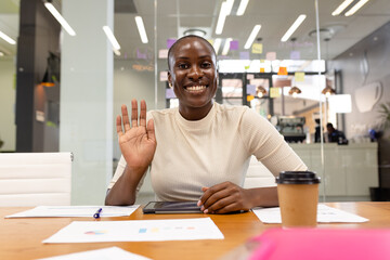 Portrait of smiling african american female professional with reports on desk waving hand in office