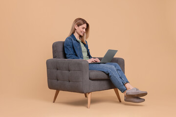 Happy woman with laptop sitting in armchair on beige background