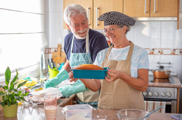 Happy senior couple in the home kitchen while wife holds a freshly baked homemade plumcake and husband washes dishes