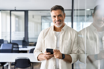 Smiling mature Latin or Indian businessman holding smartphone, using cellphone mobile app. Middle aged hispanic employee standing with phone and looking at camera on office background with copy space.