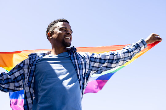 Cheerful african american young gay man with rainbow flag stretching arms against clear sky