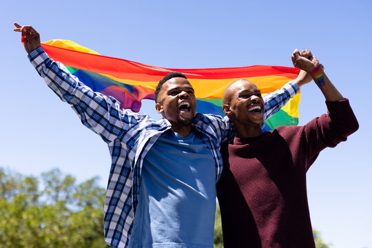 Cheerful african american gay couple with rainbow flag stretching arms under clear sky on sunny day
