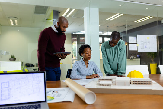 African american male and female coworkers discussing blueprint and model at desk in office
