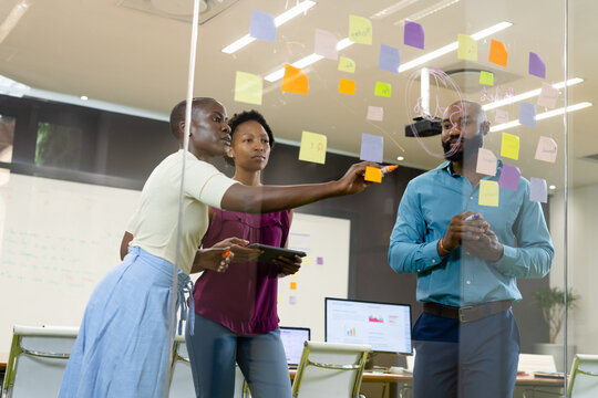 African american coworkers writing on glass wall and discussing over adhesive notes in office