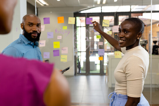 African american colleagues discussing strategies over adhesive notes on glass wall in office