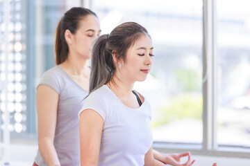 Fototapeta premium Two people Asian beautiful healthy fit slim female sporty athlete model in sport bra and leggings sitting crossed legs in lotus posture on yoga carpet smiling doing meditation in living room at home