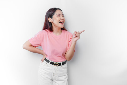 Excited Asian Woman Is Wearing Pink T-shirt, Pointing At The Copy Space Beside Her, Isolated By White Background